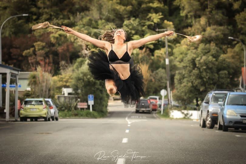 Alexandra leaping with her arms extended on a tree-lined street.