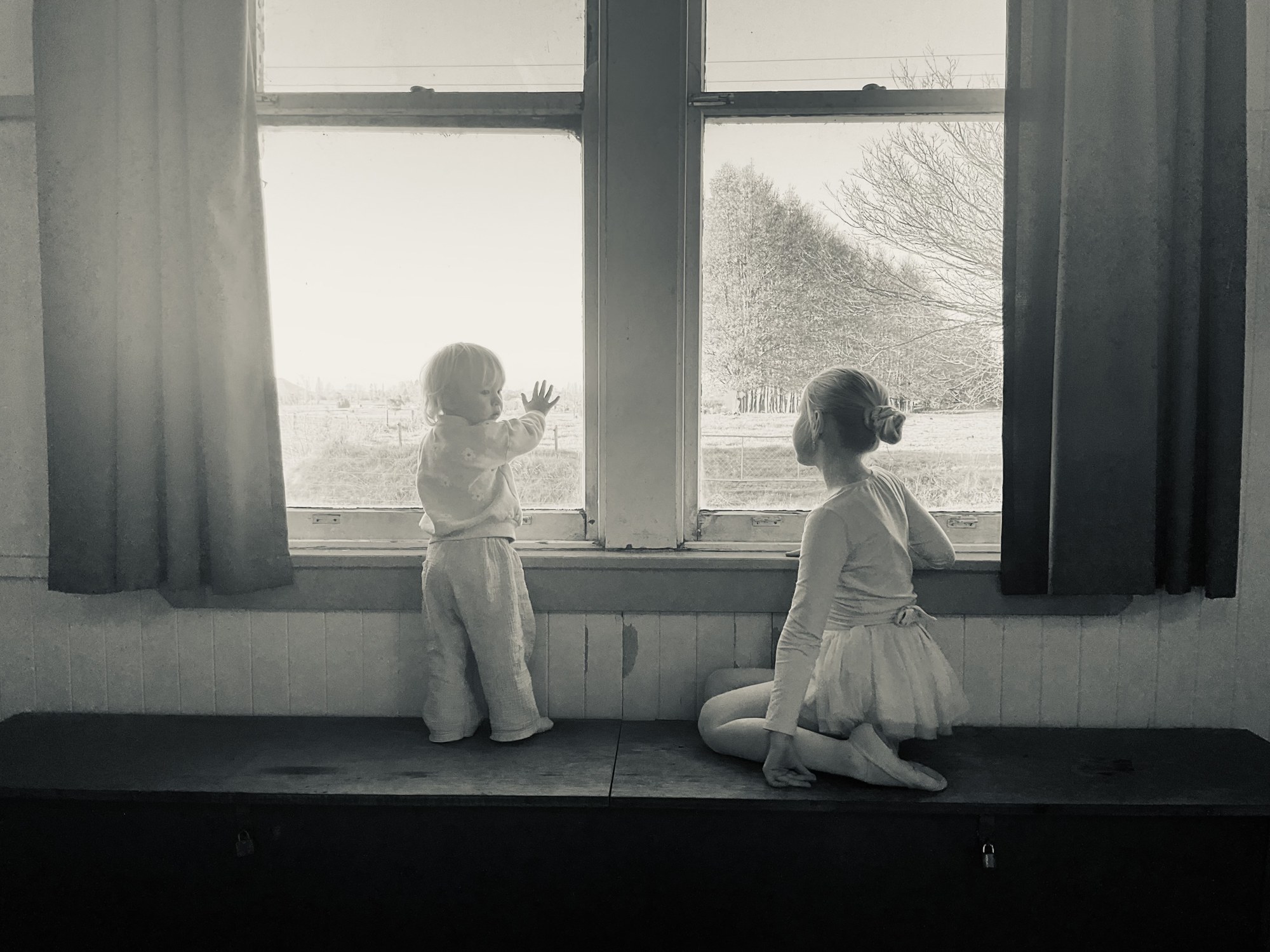 Two young children in ballet clothes looking through a sunlit hall window.